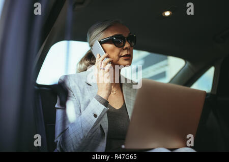 Senior Geschäftsfrau zu Office in einem Auto saß auf dem Rücksitz mit Laptop und Gespräch am Handy reisen. Geschäftsfrau mit Laptop empfangen einer Ph Stockfoto