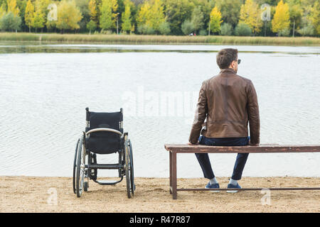 Ein junger Mann sitzt auf einer Bank am See, neben seinem Rollstuhl. Stockfoto