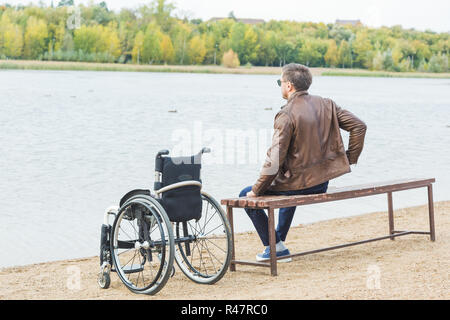 Ein junger Mann sitzt auf einer Bank am See, neben seinem Rollstuhl. Stockfoto