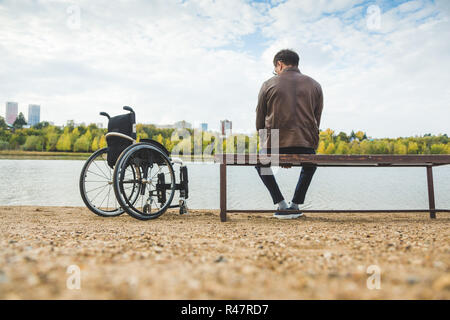Ein junger Mann sitzt auf einer Bank am See, neben seinem Rollstuhl. Stockfoto