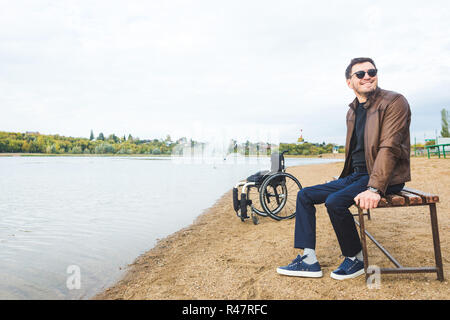 Ein junger Mann sitzt auf einer Bank am See, neben seinem Rollstuhl. Stockfoto