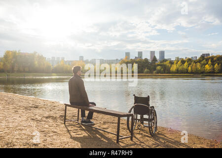 Ein junger Mann sitzt auf einer Bank am See, neben seinem Rollstuhl. Stockfoto