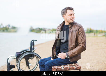 Ein junger Mann sitzt auf einer Bank am See, neben seinem Rollstuhl. Stockfoto