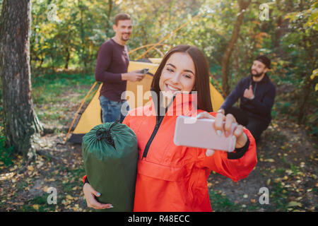 Bild der schönen jungen Frau nimmt selfie und lächelt. Sie hält Schlafsack. Junge Männer auf den Rücken wirft. Sie arbeiten mit Zelt. Stockfoto