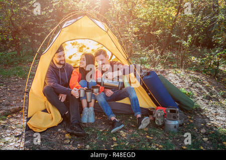 Fröhliche Freunde sitzen im Zelt und schauen sich an. Sie lächeln. Junge Frau hat Cup in den Händen. Mann auf der rechten gießt wie Trinken in den Becher. Stockfoto