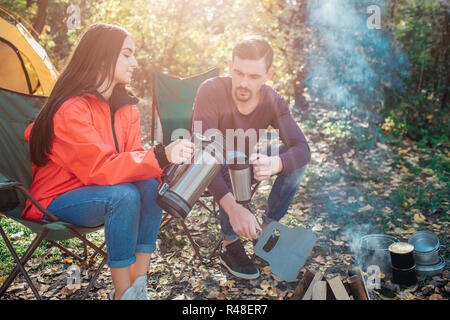 Junge Frau sitzen auf Klappstuhl und poures heißes Wasser aus Thermoskanne in man's Cup. Sie hat es genau. Kerl halten Thermotasse in der Hand. Sie sitzen am Feuer zusammen. Stockfoto