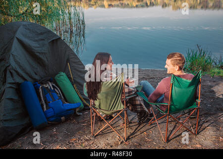 Schöne Paar zusammen sitzen auf Klappstühlen am Wasser und schauen sich an. Sie lächeln. Sie hält Schale in den Händen und mit Decke abgedeckt. Es gibt Zelt und Ausrüstung. Stockfoto