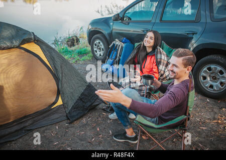 Fröhlicher junger Mann auf der Suche nach Links und die Hand. Frau schaut in die gleiche Richtung. Sie haben thermocups in Händen. Menschen sitzt am Zelt mit Auto. Stockfoto