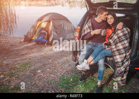 Junge Mann Frau umarmen. Sie sitzen im Kofferraum. Modell ist mit Decke abgedeckt. Paar ist am See. Es gibt Zelt an der Wasserlinie. Stockfoto