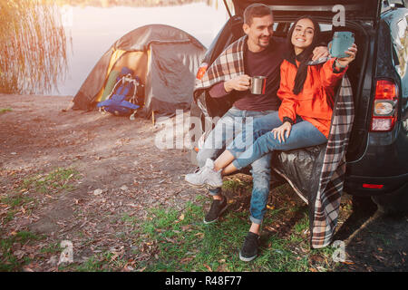 Junge Frau sitzt im Kofferraum mit Mann und vor der Kamera posieren. Sie nimmt selfie. Junge Frau halten Sie es in der Hand. Kerl hat Cup. Sind Sie am See. Stockfoto