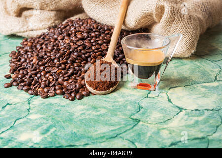 Kaffeebohnen in Kaffee sackleinen Beutel auf grüne Oberfläche, Löffel aus Holz mit gemahlenen Kaffee auf und Kaffee Glas Schale. Stockfoto