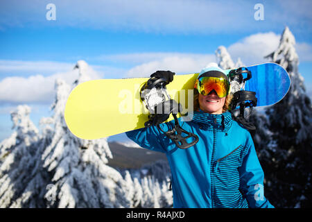Lächelnd posiert mit Snowboard Snowboarder auf der Schulter am Ski Resort in der Nähe von Forest vor backcountry Freeride und Tragekomfort Reflektierende Schutzbrille, bunte Mode Outfit. Moderne Snowboardausrüstung. Stockfoto