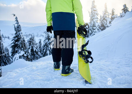 Rückansicht der Snowboarder Klettern mit seinem Brett auf dem Berg für backcountry Freeride Session im Wald. Mann mit Snowboard Wandern am Ski Resort. Reiter lime modische Outfit. Stockfoto