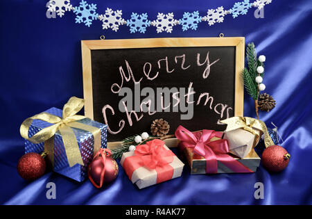 Gruß an Weihnachten, am schwarzen Brett um geschenkboxen und immergrünen Dekorationen auf blauer Seide Hintergrund Stockfoto