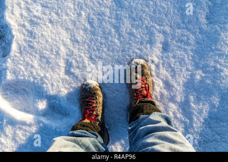 Aus erster-Person-Sicht einer Person mit Wanderschuhen und Jeans, die bei hellem Sonnenlicht auf einem frisch verschneiten Feld steht Stockfoto