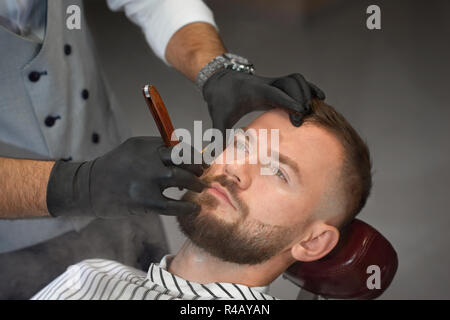 Blick von oben Der überzeugten Mann auf Stuhl in der Friseur- und Suchen während der professionellen Friseur in schwarz Handschuhe halten und vorsichtig rasieren Rasiermesser Bart von männlichen Klienten. Stockfoto