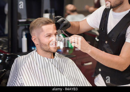 Lächelnd Mann sitzt auf Stuhl in Friseur beim Friseur in Schutzhandschuhe Entfernen von schwarzen Maske vom Gesicht des Client. Schöner Mann, Reinigung Verfahren gegen Punkte. Konzept der Pflege. Stockfoto