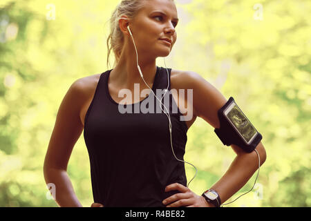 Passen junge blonde Frau in Sportkleidung Wiedergabe von Musik auf einem Handy in ein Armband beim Joggen durch den Wald Stockfoto