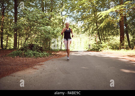 Passen junge blonde Frau in Shorts und einem Tanktop entlang einem Pfad durch einen Wald an einem sonnigen Tag Stockfoto