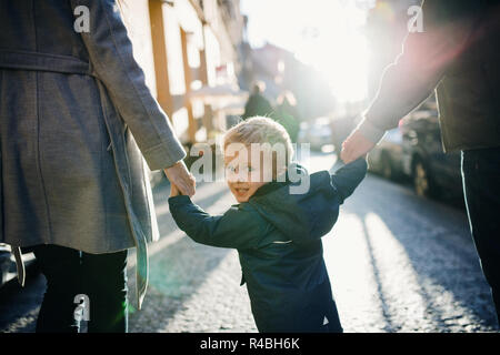 Eine Ansicht der Rückseite kleine Kleinkind Junge mit unkenntlich Eltern gehen im Freien in der Stadt. Stockfoto
