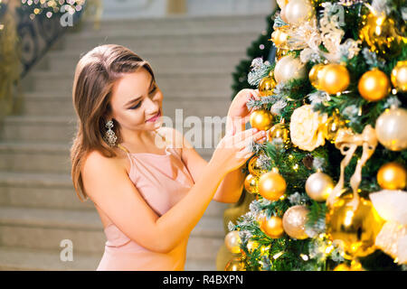 Schöne junge Frau in einem rosa elegantes Abendkleid in einem eleganten Halle und Dressing Weihnachtsbaum mit gelben Kugel auf einem Silvester. Stockfoto