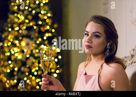 Portrait von eine schöne junge Frau sitzt in einem dunklen chic Saal mit einem Weihnachtsbaum in einer Girlande in ampir Stuhl mit einem Glas Champa gekleidet Stockfoto