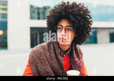 Junge curly Mixed-race Junge trinkt Kaffee auf der Straße vor dem Hintergrund von Bürogebäuden. Jugend Kultur und Mode. Student auf eine Pause zwisch Stockfoto