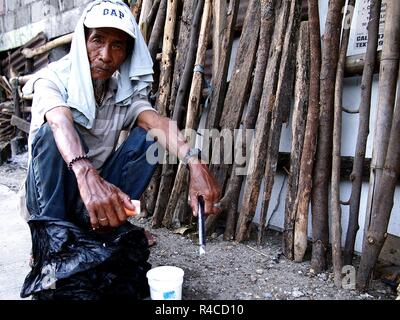 ANTIPOLO CITY, Philippinen - 24. NOVEMBER 2018: Eine alte asiatische Mann ruht, schaut die Kamera auf einem Bürgersteig entlang einer befahrenen Straße. Stockfoto
