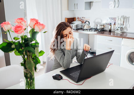Frau Arbeiten am Laptop auf moderne Küche. Junge müde Freiberufler nimmt Brille wegen Schmerzen am Auge. Stockfoto