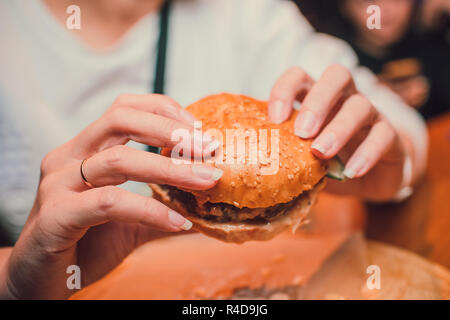 Mädchen, dass eine Gebissen ein Stück Burger in einer Hand. Mädchen mit lecker lecker fast food Cheeseburger. Das Konzept des Fast Food. Lecker ungesund Burger Sandwich in den Händen, Stockfoto