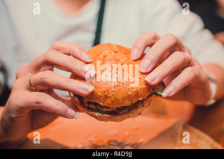 Mädchen, dass eine Gebissen ein Stück Burger in einer Hand. Mädchen mit lecker lecker fast food Cheeseburger. Das Konzept des Fast Food. Lecker ungesund Burger Sandwich in den Händen, Stockfoto