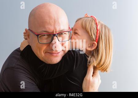 Süße liebevolle kleine blonde Mädchen küssen ihr Papa als Er umarmt sie eng in seine Arme, während in die Kamera schaut mit einem erfreuten Lächeln Stockfoto