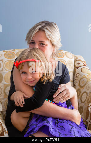 Liebevolle Mutter umarmte ihre süsse kleine Tochter, als sie sitzt an der Kamera zu lächeln, während sie sich in einen bequemen Sessel zu Hause Stockfoto