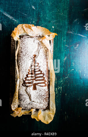 Bananenbrot, verziert mit einer weißen Weihnachtsbaum auf der Oberseite über eine rustikale grün und blau hintergrund stenciled Schokolade, Ansicht von oben. Stockfoto