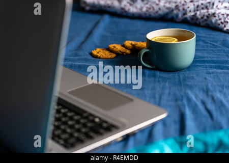 Tee mit Zitrone und Kekse mit Schokolade Verlegung auf Matratze - grau Laptop daneben, verschwommenes Türkis Hintergrund Stockfoto