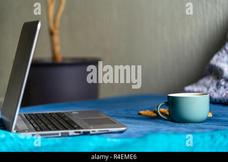 Tee mit Zitrone und Kekse mit Schokolade Verlegung auf Matratze - grau Laptop daneben, verschwommenes Türkis Hintergrund Stockfoto