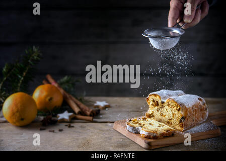 Verschwommen sieben Puderzucker auf einem Weihnachtskuchen, in Deutschland Christstollen, orange und Gewürze in die Rückseite auf einem rustikalen Holztisch, dunklen Hintergrund Stockfoto