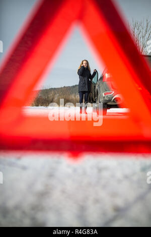 Junge Frau ein Warndreieck Einrichtung und Unterstützung gefordert, nachdem ihr Auto in der Mitte des nirgendwo an einem eiskalten Wintertag ist kaputt Stockfoto