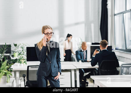 Ernsthafte junge geschäftsfrau sprechen von Smartphone, während Kollegen in Open Space Office Stockfoto