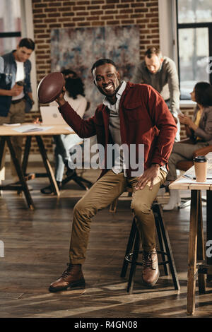 Lächelnd african american and Geschäftsmann Holding rugby ball mit Kollegen hinter Arbeiten im Loft Büro Stockfoto