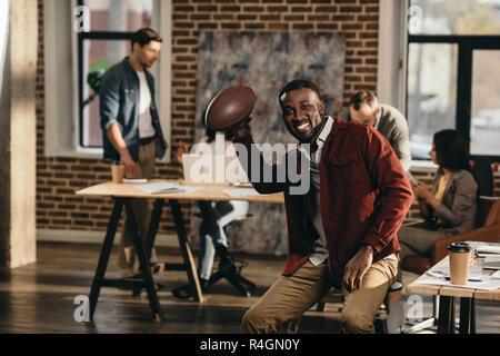 Lächelnd african american and Geschäftsmann Holding rugby ball mit Kollegen hinter Arbeiten im Loft Büro Stockfoto