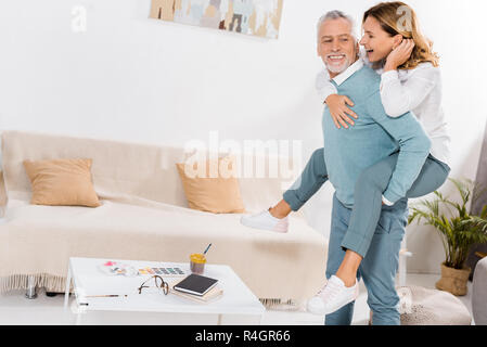 Glücklich im mittleren Alter Mann tun piggyback Ride schöne Frau im Wohnzimmer zu Hause Stockfoto