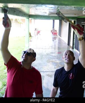 AGAT, Guam - September 27, 2018 - U.S. Navy Aviation Ordnanceman 1. Klasse Marcos Hernandez von San Antonio, Texas, Links, und US-Navy Aviation Ordnanceman 3. Klasse David Nguyen kratzen den Lack von der Obergrenze der Bürokomplex der Bürgermeister mit Agat Agat Mitarbeiter in der Nähe. U.S. Navy Matrosen von Naval Base Guam und USS Ronald Reagan und US Air Force Piloten von Andersen Air Force Base versammelten sich in der Weinlaube, Guams Bürgermeister Büro heute für ein Dorf zu reinigen. Die Gruppe von fast 40 service Mitglieder aufgeteilt in zwei Gruppen. Die ersten versammelt 2 Muldenkipper voller Vegetation, von denen viel war Klopfen Stockfoto