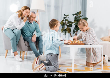 Lächelnde Kinder Schach spielen, während ihre Großeltern in der Nähe von zu Hause sitzen Stockfoto