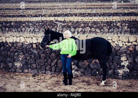 Fashion Concept mit cute blonde Model posiert in der Landschaft mit einem schwarzen Pferd und das Tragen einer grünen Jacke. Farben und Kontrast, um Menschen und Tiere Stockfoto