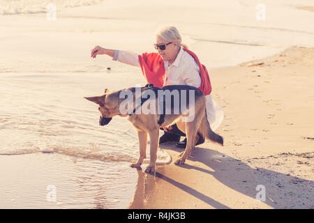 Schönen Witwe ältere Frau mit ihr spielen Deutscher Schäferhund Hund genießen ihre Begleiter und Liebe am Strand bei Sonnenuntergang in Vorteile von Tieren acti Halten Stockfoto