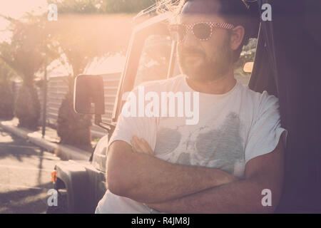 Mann mittleren Alters mit Sonnenbrille in der nähe von Off road Auto in tropischen Straße mit Sonne im Gegenlicht geparkt - Sonnenuntergang Atmosphäre für schöne kaukasischen Mitarbeiter Stockfoto