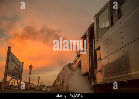 Niedriger Winkel, Seitenansicht Nahaufnahme des alten britischen Dampfzugs am Bahnsteig in leerem historischen Bahnhof am Ende des Tages mit glühendem Sonnenuntergang Abendhimmel. Stockfoto