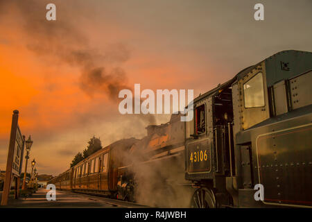 Niedriger Winkel, Seitenansicht Nahaufnahme des alten britischen Dampfzugs am Bahnsteig in leerem historischen Bahnhof am Ende des Tages mit glühendem Sonnenuntergang Abendhimmel. Stockfoto