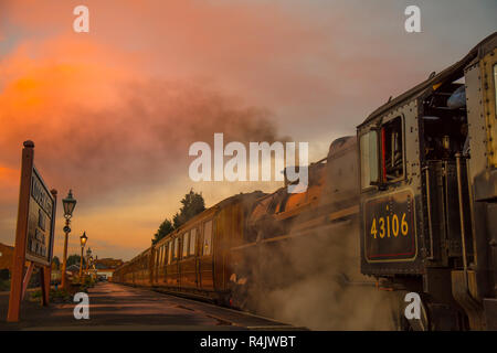 Niedriger Winkel, Seitenansicht Nahaufnahme des alten britischen Dampfzugs am Bahnsteig in leerem historischen Bahnhof am Ende des Tages mit glühendem Sonnenuntergang Abendhimmel. Stockfoto